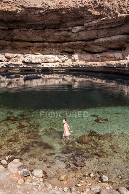 Расслабленная азиатская женщина, оглядываясь назад на камеру на прозрачной воде Bimmah Sinkhole окруженный грубыми камнями во время путешествия в Оман — стоковое фото