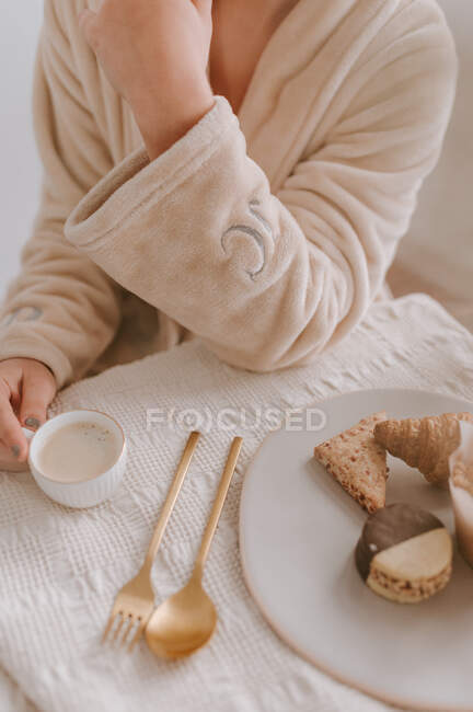 Сверху урожая анонимная женщина в халате сидит за столом с чашкой кофе и сладкими десертами во время завтрака дома — стоковое фото