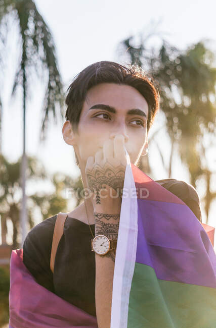 Tranquilo hombre gay con tatuajes envueltos en colorida bandera LGBT en la calle de la ciudad mirando hacia otro lado - foto de stock