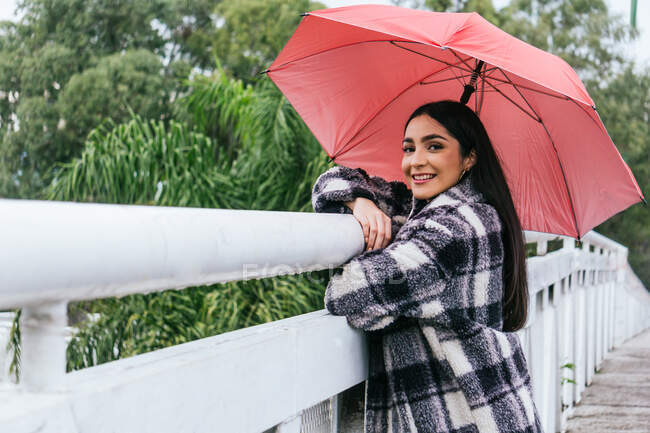 Vista laterale di femmina etnica ottimista con ombrello sorridente e guardando la fotocamera mentre si appoggia sulla ringhiera del ponte nella giornata piovosa nel parco — Foto stock