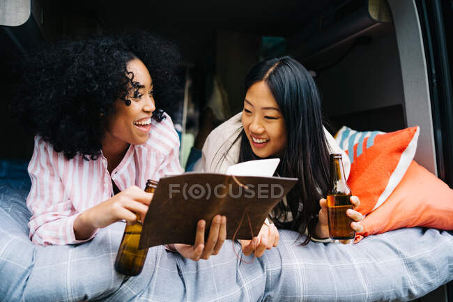Cheerful young multiracial women drinking beer and discussing travel route with map while chilling together in camper van during summer journey — Stock Photo