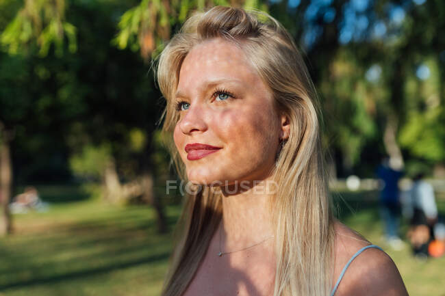 Happy female with blond hair and shadow on face standing in summer park on sunny day and looking away — Stock Photo