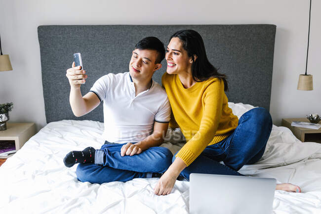 Восхитительная этническая мать и мальчик-подросток с синдромом Дауна сидят на кровати и делают автопортрет на смартфоне — стоковое фото