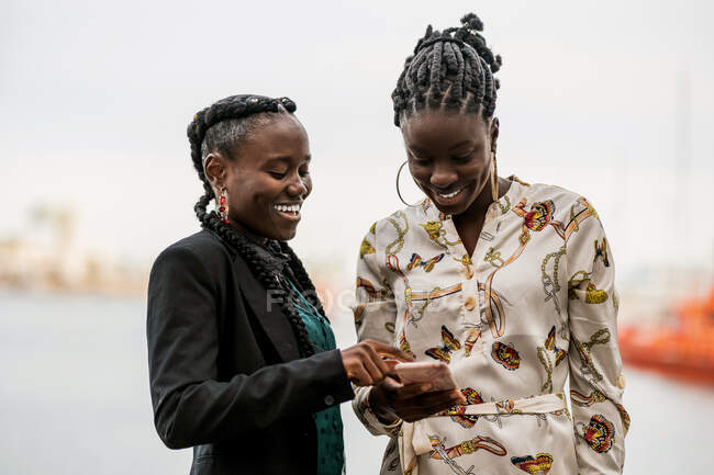 Las damas afroamericanas sonrientes de moda con peinado pasan tiempo juntos navegando por el teléfono móvil en el parque en un día brillante - foto de stock