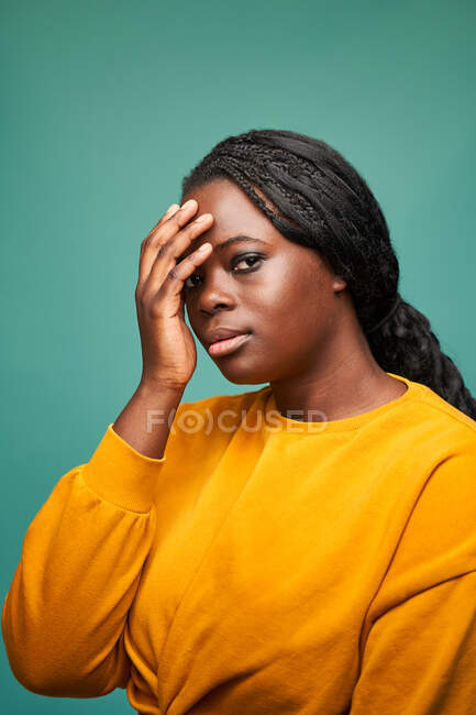 Афроамериканська жінка в жовтому светрі і кашкет торкається обличчя і дивиться на камеру, стоячи проти синьої стіни. — стокове фото