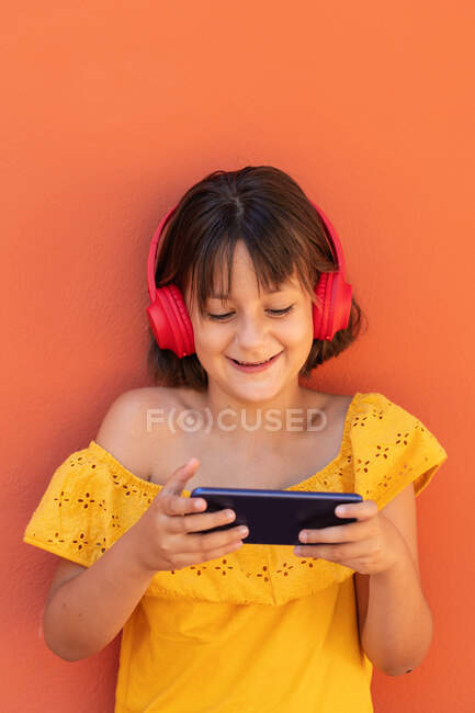 Inhalt Kind surft mit dem Handy im Internet, während es Songs vom drahtlosen Headset auf orangefarbenem Hintergrund hört — Stockfoto