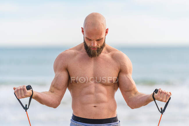 Shirtless brutal man looking away doing side lateral flies with resistant band training on seashore — Stock Photo