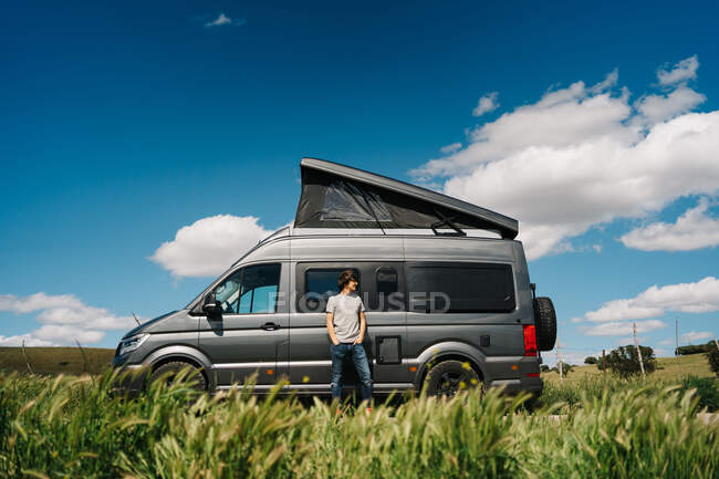 Полное тело молодого авантюриста смотрит в сторону, когда отдыхает возле фургона с палаткой, припаркованной в летней сельской местности. — стоковое фото