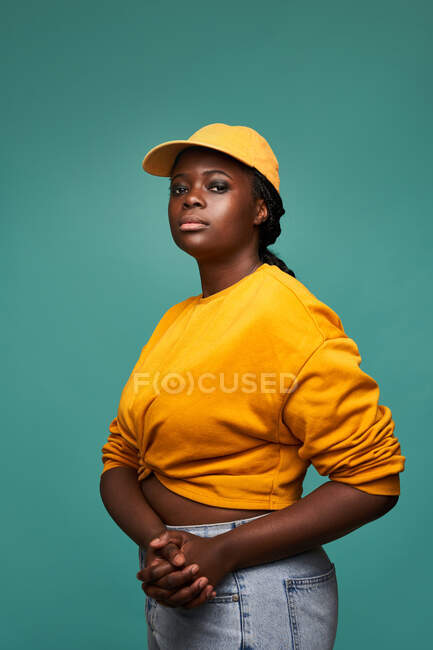 Unemotional grassoccio afroamericano femminile in maglione giallo e cappuccio con le mani insieme guardando la fotocamera mentre in piedi contro il muro blu — Foto stock
