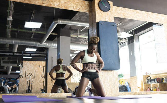 Athlète afro-américaine en vêtements de sport avec les mains sur les hanches respirant pendant l'entraînement contre miroir dans le gymnase — Photo de stock