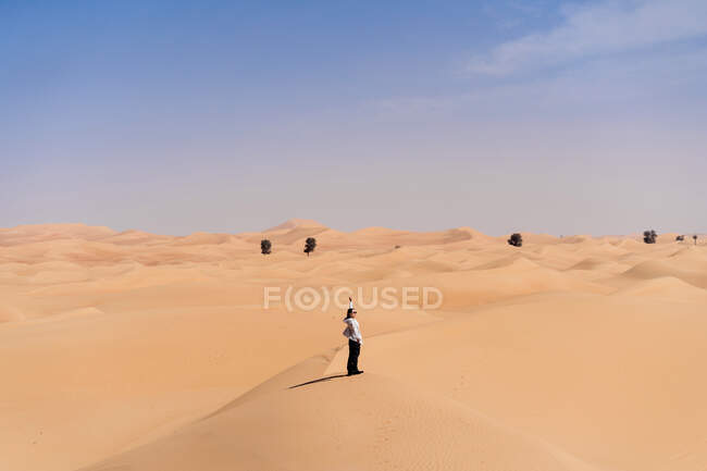 Lächelnde junge Frau in lässiger Kleidung, die während einer Reise in den Emiraten auf einer Sanddüne in der Wüste steht und die Arme ausstreckt — Stockfoto