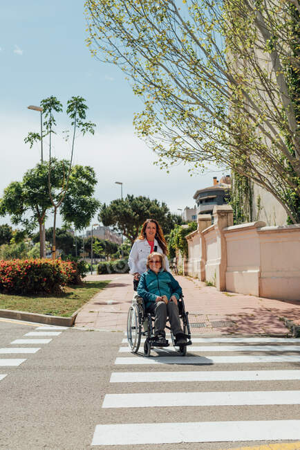 Взрослая женщина толкает инвалидное кресло со старшей матерью и пересекает дорогу в городе во время прогулки летом — стоковое фото