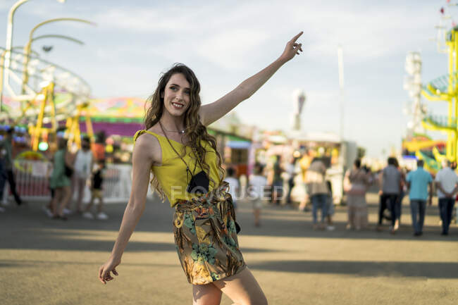 Восхитительная женщина, стоящая перед камерой с поднятыми руками на ярмарке и наслаждающаяся летними выходными — стоковое фото