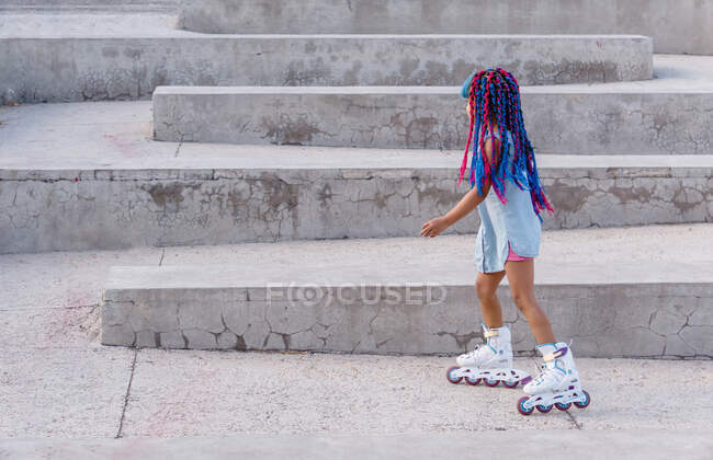 Vue latérale de l'enfant ethnique en patins à roulettes avec des tresses colorées debout sur l'escalier — Photo de stock