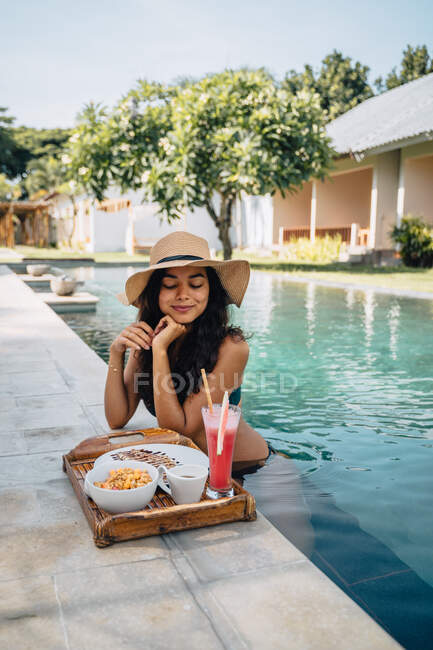 Touriste féminine joyeuse penchée au bord de la piscine tout en regardant vers le bas contre plateau avec un délicieux petit déjeuner au soleil — Photo de stock