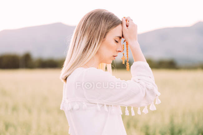 Vista lateral de mujer fiel en vestido blanco sosteniendo cuentas con cruz mientras que da oraciones en soledad en el campo rural tranquilo en la naturaleza - foto de stock