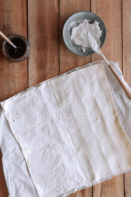Vista superior del frasco con mermelada de higo y un tazón con queso crema colocado cerca de la masa de pastelería fina en la mesa de madera - foto de stock