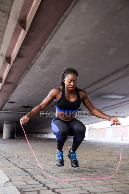 Donna afro-americana in abbigliamento sportivo tenendo la corda di salto e guardando la macchina fotografica mentre in piedi sul marciapiede sulla strada della città — Foto stock