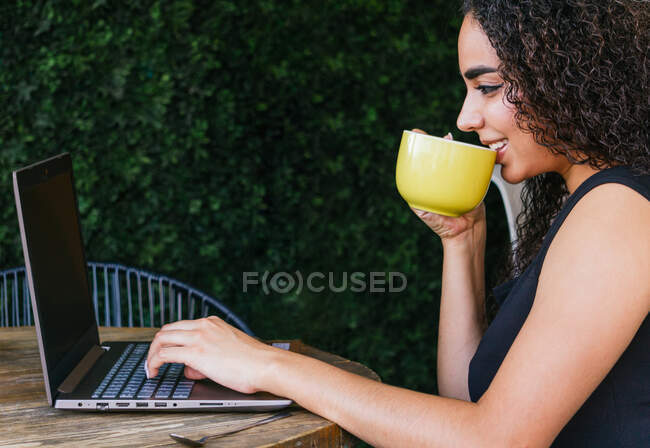 Vista lateral del contenido joven freelancer étnico bebiendo café y escribiendo en el teclado del ordenador portátil mientras trabaja en un proyecto remoto en la terraza de la cafetería - foto de stock