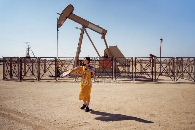 Mujer joven de cuerpo entero en vestido tradicional de pie sobre tierra arenosa contra plataforma de perforación de petróleo durante su visita al campo petrolífero en Bahréin - foto de stock