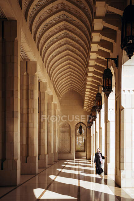 Полная длина неузнаваемой женщины в традиционной одежде, идущей по декоративной арке Большой мечети Султана Кабуса в Маскате — стоковое фото