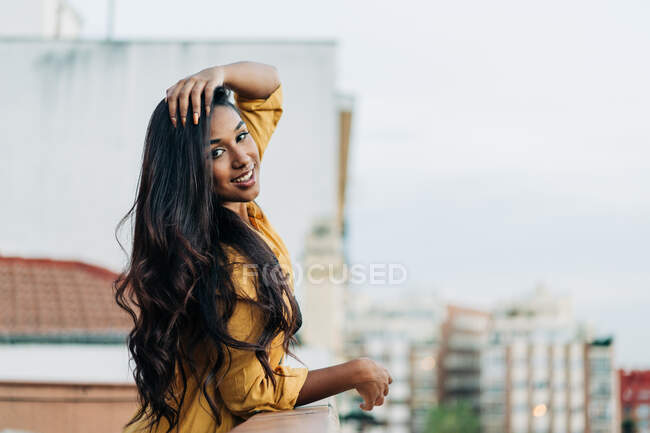 Young delighted Hispanic female in casual clothes leaning on railing and looking at camera while relaxing on balcony in evening in city — Stock Photo
