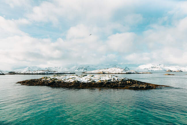Steinige Inselchen im plätschernden Meer in der Nähe des schneebedeckten Bergrückens vor bewölktem Himmel im Winter auf den Lofoten, Norwegen — Stockfoto
