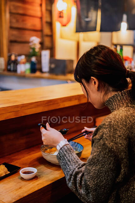 Vista lateral da mulher asiática satisfeita em suéter sorrindo enquanto toma colher do trabalhador sentado no balcão de madeira no bar ramen — Fotografia de Stock