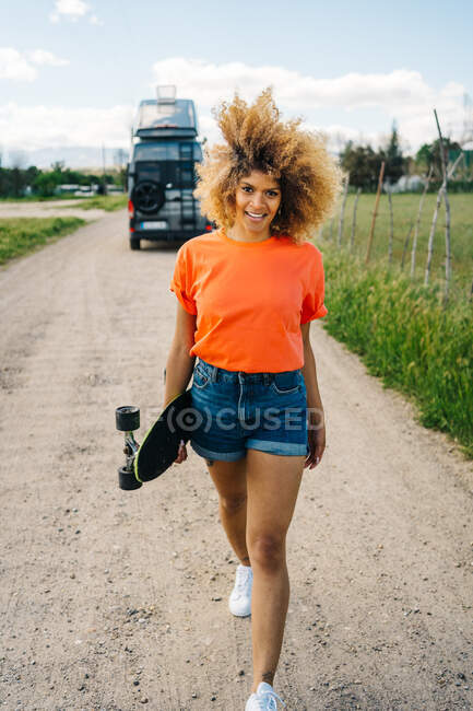 Felice donna afroamericana che porta il longboard e distoglie lo sguardo con il sorriso mentre cammina sulla strada di campagna vicino al furgone in estate — Foto stock