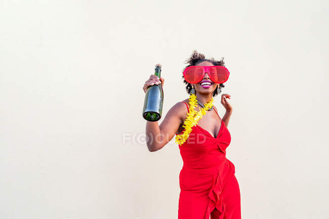Cheerful African American female in red apparel and glasses holding bottle of alcoholic beverage during party on light background — Stock Photo