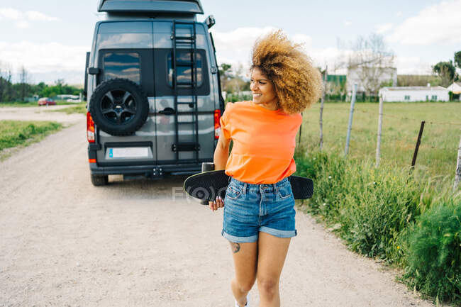 Счастливая афроамериканка носит лонгборд и с улыбкой смотрит в сторону, когда идет по сельской дороге возле фургона летом — стоковое фото