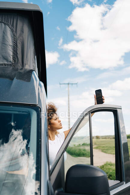 Infeliz joven afroamericana mujer de pie en la puerta del conductor en autocaravana y tratando de obtener la conexión en el teléfono móvil, mientras que tiene problemas durante el viaje por carretera - foto de stock