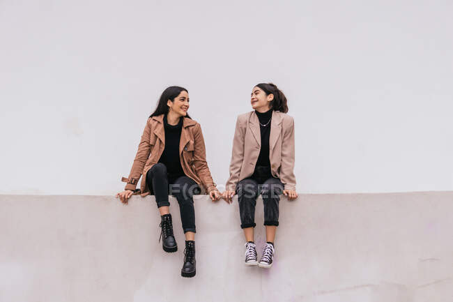 Full body cheerful ethnic young women smiling and sitting on gray wall on city street — Stock Photo