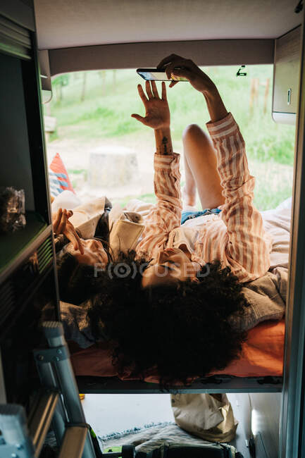 Jóvenes novias multirraciales tumbadas juntas en autocaravana y tomando selfie en el teléfono inteligente mientras se enfría y disfruta de las vacaciones de verano en la naturaleza - foto de stock