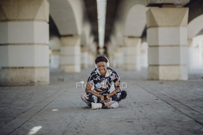 Jovem aptidão afro-americana mulher descansando e esticando as pernas enquanto ouve música depois de correr na rua da cidade — Fotografia de Stock