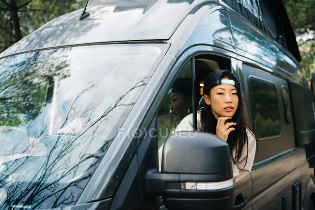 Heureux jeune voyageuse asiatique en chapeau regardant par la fenêtre ouverte du camping-car et profitant de la liberté pendant le voyage à travers la campagne estivale — Photo de stock