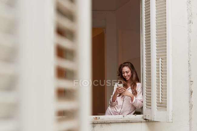 Cheerful female in pajama standing near window and browsing mobile phone at home — Stock Photo