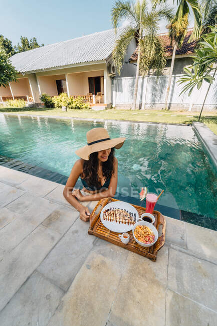 Turista feminino alegre inclinado na beira da piscina enquanto olha para baixo contra bandeja com delicioso café da manhã à luz do sol — Fotografia de Stock