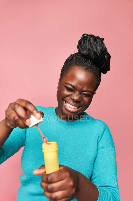 Happy African American female with eyes closed wearing blue clothes and blowing soap bubbles against pink background in studio — Stock Photo