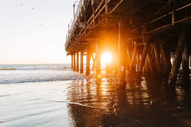 Glowing sunlight at evening time penetrating piles of Santa Monica pier with peaceful ocean waves running on beach in California — Stock Photo