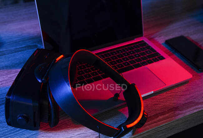 Alto ángulo de escritorio de madera con netbook abierto cerca del teléfono y gafas VR en habitación oscura con luces de neón - foto de stock