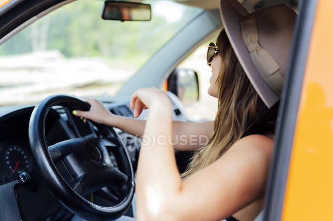 Vista lateral de la viajera sentada en el asiento del conductor en furgoneta y disfrutando del viaje por carretera en verano - foto de stock