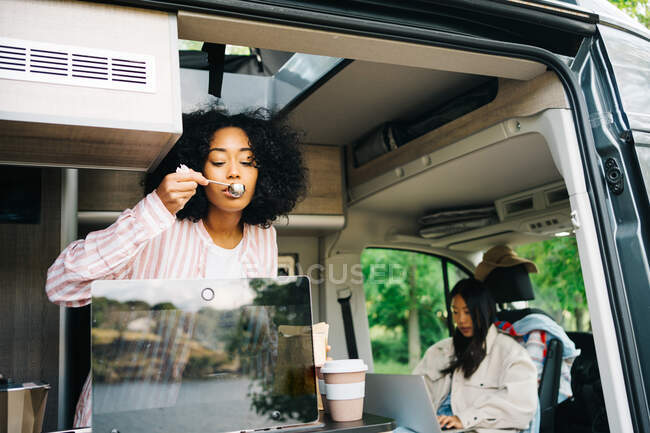 Joven mujer negra preparando café mientras ve vídeo en el ordenador portátil dentro del vehículo campista con la novia asiática durante el viaje de verano en la naturaleza - foto de stock