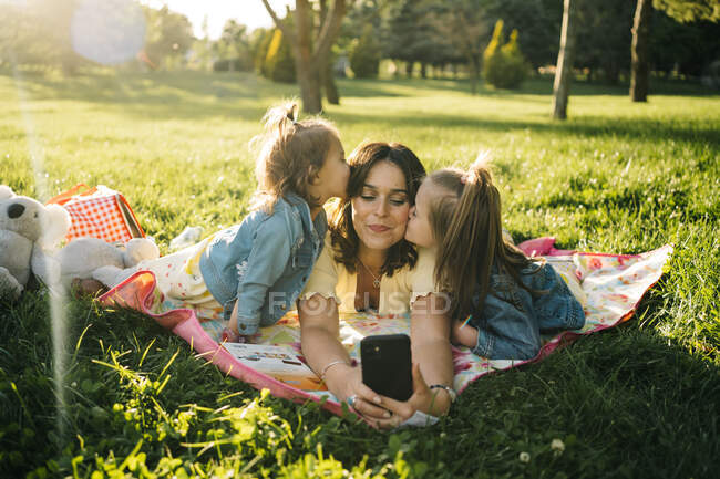 Счастливая молодая женщина и милые маленькие дочери лежат на одеяле и делают селфи на смартфоне, веселясь вместе на зеленом лугу в летнем парке — стоковое фото