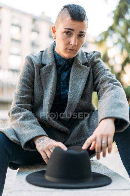 Queer in stylish apparel and hat with tattoo and mohawk looking at camera while sitting in town — Stock Photo