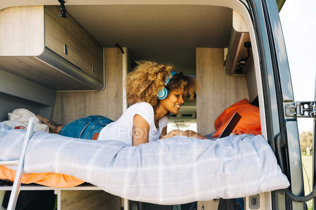 Веселая афроамериканка улыбается и слушает музыку в наушниках, лежа на кровати в фургоне и просматривая социальные сети на ноутбуке — стоковое фото