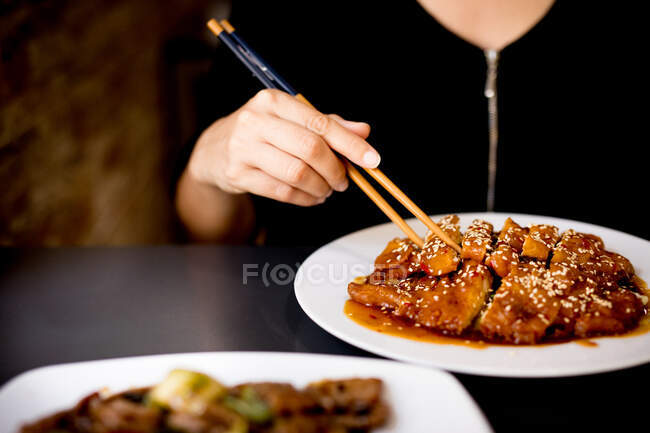 Cultiver femelle en robe élégante assis dans un restaurant asiatique et manger préparé poulet épicé savoureux avec baguettes — Photo de stock