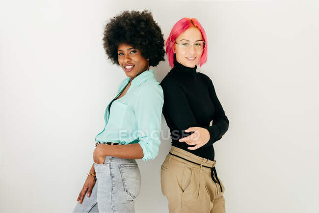 Fröhliche junge Frau mit rosa Haaren und afroamerikanische Freundin in stilvollem Outfit stehen Rücken an Rücken auf weißem Hintergrund — Stockfoto