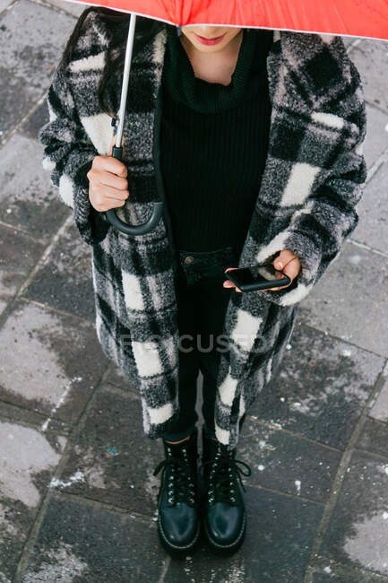 De cima jovem fêmea étnica em checkered casaco de navegação celular enquanto de pé sob guarda-chuva vermelho no dia chuvoso na rua molhada — Fotografia de Stock