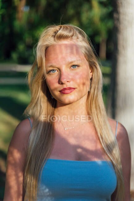 Glückliches Weibchen mit blonden Haaren und Schatten im Gesicht, das an einem sonnigen Tag im Sommerpark steht und in die Kamera blickt — Stockfoto